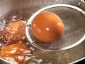 Jak ugotować jajka? Brązowe jajka gotują się w bulgoczącej wodzie, jedno z nich jest wyjmowane sitkiem.