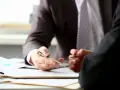 Dwaj mężczyźni w garniturach omawiają dokumenty. Jeden z nich wskazuje piórem na umowę, co może symbolizować niemiecki dług i jego egzekwowanie w Polsce.