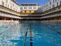 Piscine art déco parisienne, avec ses bâtiments jaunes et blancs, ses balcons et ses transats. L'eau bleue invite à la baignade.