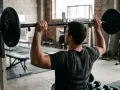 Un homme effectue des exercices de musculation avec une barre chargée dans une salle de sport rustique.