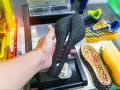 A hand holds a black, intricately patterned bicycle saddle, likely made from tpu material. In the background, colorful 3D-printed shoe soles and a shoe are visible.