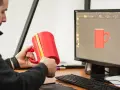 Mężczyzna mierzy czerwoną kubek 3d, porównując go z modelem na ekranie komputera.