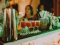 Cztery kieliszki z czerwonym drinkiem stoją na barze, gotowe do podania na weselu. W tle widać gości i dekoracje.