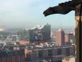 Blick über Hamburg mit der Elbphilharmonie, gesehen durch ein Fenster des Mahnmals St. Nikolai. Ein Wasserspeier wacht über die Stadt.