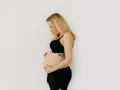 Kobieta w czarnym biustonoszu i spodenkach delikatnie obejmuje swój brzuch w 3 miesiącu ciąży.
