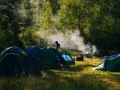 Słoneczny poranek na polu biwakowym. Kilka namiotów rozbitych wśród drzew, unosi się dym z ogniska.