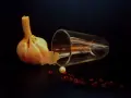 Photographie nature morte : une tête d'ail, un verre renversé, une perle et des baies de poivre rouge sur fond sombre.