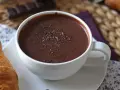 En vit mugg med varm choklad, toppad med chokladspån. En croissant ligger bredvid.