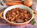Piatto fumante di salsiccia e fagioli in umido, con carote, cipolla e aglio sullo sfondo.