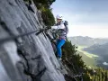 Kobieta w kasku i uprzęży pokonuje via ferrata, chwytając się stalowej liny. W tle rozciąga się malownicza górska dolina.