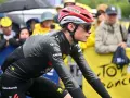 Trine Vingegaard, a cyclist in a grey and black racing suit, rides in the rain. Spectators watch from behind a yellow barrier with "Tour de France" branding.