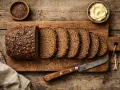 Ein Laib dunkles, kerniges Schwarzbrot, in Scheiben geschnitten, daneben Butter und Kümmelsamen. So sieht ein herzhaftes Schwarzbrot aus.