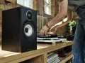 Czarny głośnik Bowers & Wilkins, jeden z najlepszych producentów głośników, obok gramofonu, na którym osoba kładzie płytę.