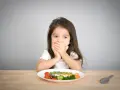 Kleines Mädchen bedeckt Mund, schaut auf Teller mit Gemüse. Manche kinder essen schnell, aber dieses hier scheint zu überlegen.