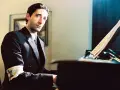 Adrien Brody jako Władysław Szpilman w filmie "Pianista" Romana Polańskiego, z opaską na ramieniu, siedzi przy fortepianie.