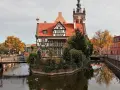 Urokliwy zakątek w **starym mieście Gdańsku**. Zabytkowa kamieniczka z czerwonym dachem i wieżą zegarową odbija się w spokojnej wodzie kanału.