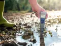 Persona con bota verde mide la calidad del agua con un medidor de pH en un entorno natural.