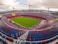 Widok na budowę stadionu, gdzie powstaje nowe Camp Nou. Żurawie górują nad trybunami w kolorach czerwonym i niebieskim, a murawa lśni zielenią.