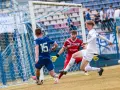 Piłkarz Michał Bartczak w białej koszulce próbuje strzelić gola, obrońca w niebieskiej koszulce z numerem 15 i bramkarz w czerwonej próbuje go zatrzymać.