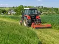 Czerwony traktor z mulczerem do c 330 kosi trawę na zielonym polu. W tle wiejskie domy i drzewa.