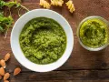 Pesto di rucola e mandorle in una ciotola bianca e un vasetto, con mandorle e rucola fresca intorno.