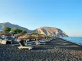 Czarne piaski i leżaki pod słomianymi parasolami na Santorini plażach. W tle góry i spokojne morze.
