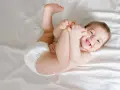 Uśmiechnięte dziecko w pieluszce leży na białej pościeli, bawiąc się swoimi stopami. To naturalne, **dlaczego dziecko liże ręce** – odkrywa świat.