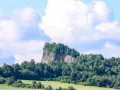 Skalisty szczyt porośnięty drzewami, przypominający wilczą górę w Złotoryi, góruje nad zielonymi polami i lasami.