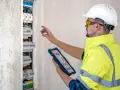 Elektryk w kasku i kamizelce odblaskowej sprawdza rozdzielnicę elektryczną na tablecie.