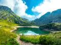 Sentiero panoramico tra le montagne, con un lago alpino dalle acque smeraldine che riflette il cielo azzurro e le nuvole bianche. Un perfetto percorso dolomiti.