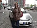 Frau mit brauner Lederjacke und roter Tasche steht neben silbernem Auto. Baustelle im Hintergrund. Sie ist free now fahrer.
