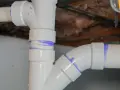 Close-up of various PVC pipe types, showing a vertical pipe connected to a horizontal pipe with a curved fitting, all marked with purple solvent cement.