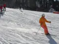 Narciarka w pomarańczowej kurtce szusuje po ośnieżonym stoku. W tle widać innych narciarzy i las. To piękne narty na Kaszubach.