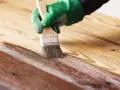 Mano con guante verde aplicando barniz a madera. Proceso para saber como barnizar madera ya barnizada y renovarla.