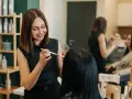Uśmiechnięta charakteryzatorka wykonuje makijaż klientce. W tle widać inne osoby pracujące w salonie.
