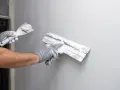 Ręce w rękawiczkach nakładają tynk na ścianę. Czas schnięcia tynku zależy od wielu czynników.
