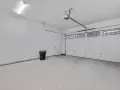 Nowy garaż z płyt betonowych projekt. Biała brama segmentowa, drzwi, czarny kosz na śmieci i sufitowe oświetlenie.