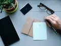 Dłoń z ołówkiem nad papierem, gotowa do napisania zwrotów do adresata. Obok notes, okulary i koperta.