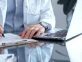 Lekarz w białym kitlu pisze na dokumentach, obok laptop. To może być zwolnienie od psychiatry.