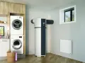 Dans une buanderie moderne, un ballon thermodynamique blanc et gris est installé près d'une fenêtre. Des machines à laver sont empilées à gauche.