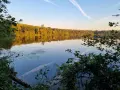Sonnenuntergang am Liblarer See. Bäume spiegeln sich im ruhigen Wasser, der Himmel ist blau mit weißen Kondensstreifen.