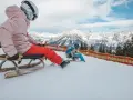 Zwei Personen rodeln auf einer verschneiten Piste in Tirol. Im Hintergrund erheben sich majestätische, schneebedeckte Berge.
