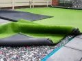Instalación de césped artificial en terraza. Se ve el rollo de césped desplegado sobre una base de goma reciclada y baldosas.
