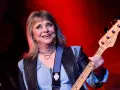 Suzi Quatro, die Rock-Ikone, spielt Bassgitarre. Sie trägt eine Lederjacke und eine Kette mit Herzanhänger. Ihr Alter ist nicht ersichtlich, aber ihre Energie ist spürbar.