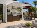 Idéal pour fabriquer un coin barbecue couvert : une cuisine extérieure moderne avec grill, évier, frigo et TV, où trois amis trinquent sous un auvent rétractable.
