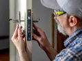 Mężczyzna w czapce i okularach ochronnych dokonuje montażu drzwi, precyzyjnie dopasowując mechanizm zamka.