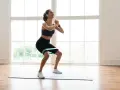 Femme souriante faisant un squat avec un élastique autour des cuisses, sur un tapis de yoga, devant une grande fenêtre lumineuse.