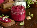 Glas mit leuchtend rotem Rote Bete Pesto, daneben Brot mit Pesto und Joghurt. Walnuss und Kräuter garnieren die Szene.