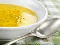 Una ciotola bianca piena di brodo di carne dorato, con goccioline d'olio in superficie. Un cucchiaio d'argento è appoggiato accanto.