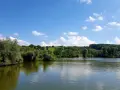 Ein sonniger Tag am Badesee Esslingen. Grüne Hügel, Bäume und ein blauer Himmel mit weißen Wolken spiegeln sich im ruhigen Wasser.